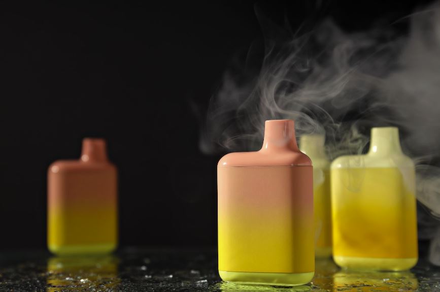 Different colored disposable vapes stand upright on a table against a black background with vapor lingering in the background.