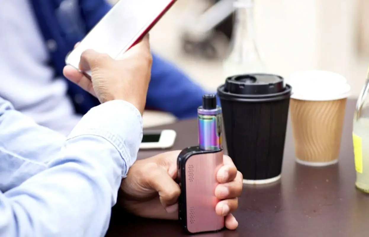 man holding a vaping device in one hand while using a cell phone next to coffee cups.