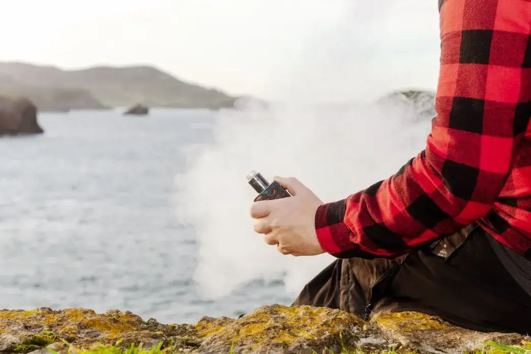 Person holding a vape and exhaling smoke outside by a body of water.