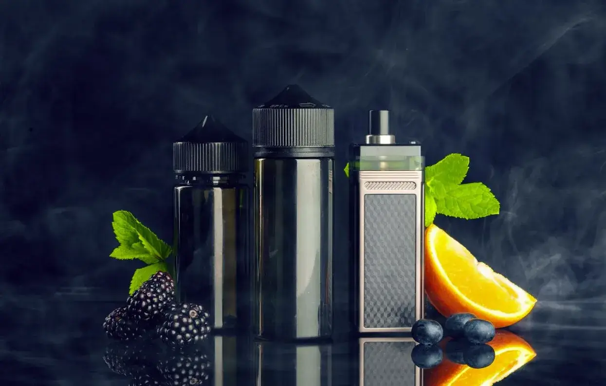 Vape device and e juice next to blackberries an orange slice and blueberries.