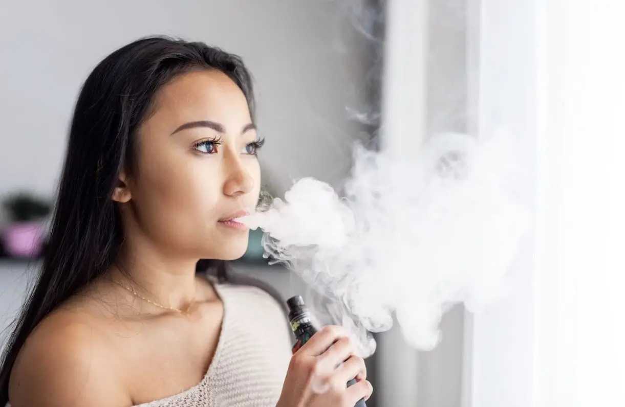 woman exhaling vapor from a vaping device next to a window and curtains.